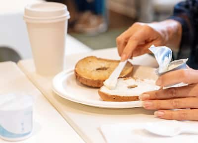 View of hands spreading cream cheese on a bagel with a cup of coffee next to the plate.