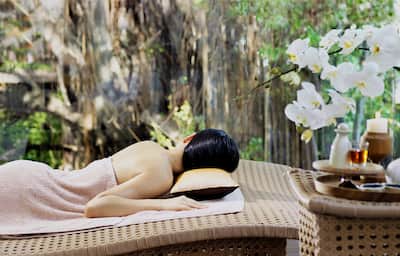 A lady lying on a message table is covered by a towel with her back to the camera. There are massage oils and flowers in the foreground and a tranquil garden in the background. 